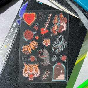 May include: A sheet of clear vinyl stickers with a variety of colorful designs, including a heart with the words "true love", a butterfly, a scorpion, a tiger, a snake, dice, and a rose.