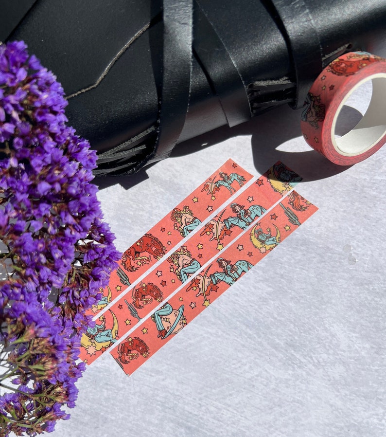 K&ouml;nnte beinhalten: Drei Rollen rosa Washi-Tape mit einem sich wiederholenden Muster von Frauen, die auf Himmelsk&ouml;rpern mit Sternen und Monden reiten. Das Klebeband befindet sich auf einer wei&szlig;en Oberfl&auml;che.