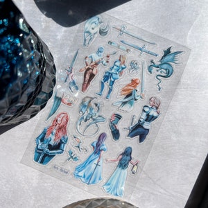 May include: A clear sticker sheet featuring fantasy-themed illustrations. The sheet includes images of knights, dragons, swords, and ethereal figures in shades of blue, red, and brown. The text "Arch Michael" is visible on the sheet.