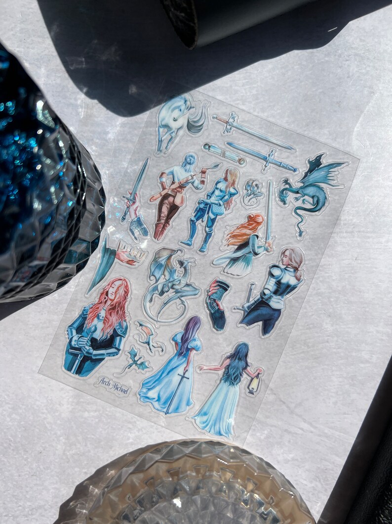 May include: A clear sticker sheet featuring fantasy-themed illustrations. The sheet includes images of knights, dragons, swords, and ethereal figures in shades of blue, red, and brown. The text "Arch Michael" is visible on the sheet.