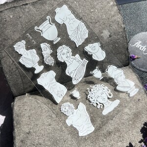 May include: A clear sheet of stickers featuring white line art illustrations of classical Greek sculptures, including busts, torsos, and vases. The stickers are arranged on a textured gray surface, with the text "Arch-Island" visible.
