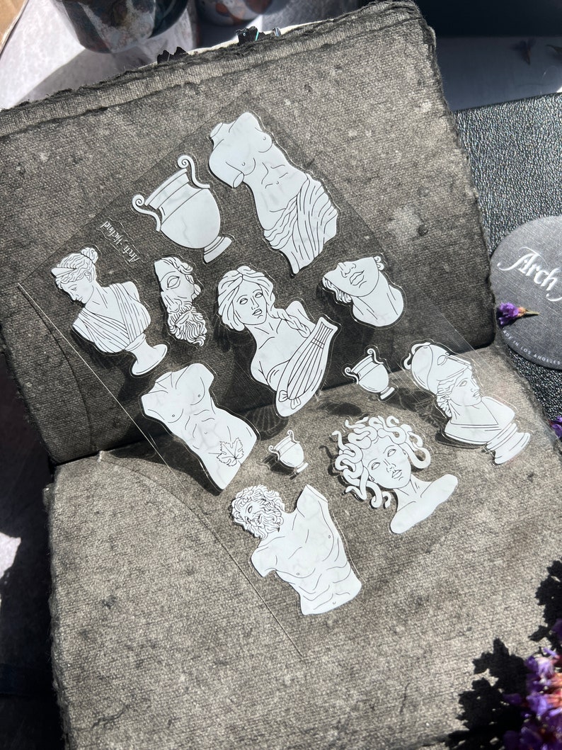 May include: A clear sheet of stickers featuring white line art illustrations of classical Greek sculptures, including busts, torsos, and vases. The stickers are arranged on a textured gray surface, with the text "Arch-Island" visible.