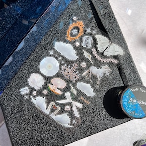 May include: A sheet of clear stickers with a white background featuring various images including swans, clouds, pearls, a castle, a seashell, a star, a diamond, a mirror, and a white horse. The stickers are on a black textured surface.