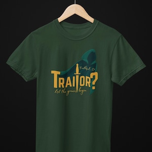 Puede incluir: Camiseta verde oscuro con la palabra "TRAITOR?" en amarillo y las palabras "Let the games begin" en cursiva. Un diseño de figura encapuchada y espada en color turquesa. La camiseta está colgada en una percha de madera.