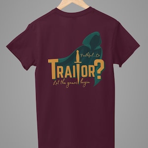 May include: Burgundy t-shirt with a gold "TRAITOR?" graphic, a green hooded cloak, and a sword design. The text "Faithful...Or" and "Let the games begin" are also included. The t-shirt is displayed on a wooden hanger.