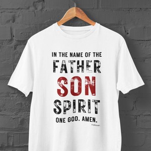 Könnte beinhalten: Weißes T-Shirt mit einem roten und schwarzen, abgenutzten Aufdruck, der "Im Namen des Vaters, des Sohnes und des Heiligen Geistes. Ein Gott. Amen." lautet.