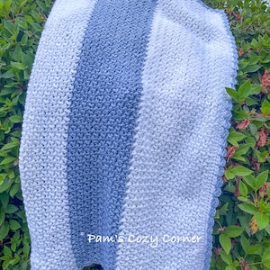 May include: A soft, knitted blanket in shades of blue and white. The blanket features vertical stripes of varying widths. The text "Pam's Cozy Corner" is visible in the lower left corner. The blanket is folded and draped over greenery.