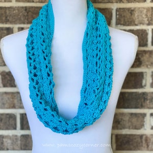 May include: A turquoise crocheted infinity scarf with a lacy pattern. The scarf is displayed on a white mannequin against a brick wall background.