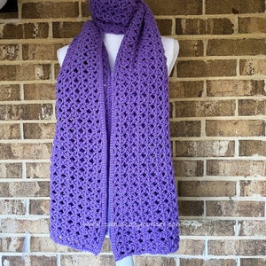 May include: A purple crocheted scarf with a lacy pattern. The scarf is displayed on a mannequin against a brick wall.