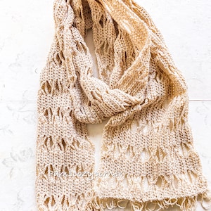 May include: A beige crocheted scarf with a lacy pattern and fringe. The scarf is folded in half and laid flat on a white surface.