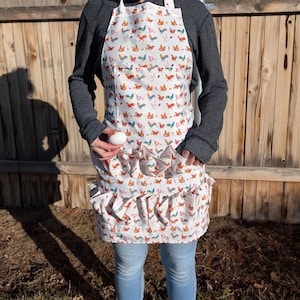 Puede incluir: Un delantal blanco con estampado de pollos coloridos y varios bolsillos. El delantal tiene un borde con volantes y lo lleva una persona que sostiene un huevo blanco.