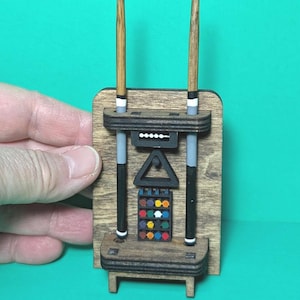 May include: A miniature wooden wall-mounted pool cue rack featuring two vertical cues with blue tips, a black triangular rack, a small abacus-style score counter with white beads, and a grid of twelve colorful miniature billiard balls.