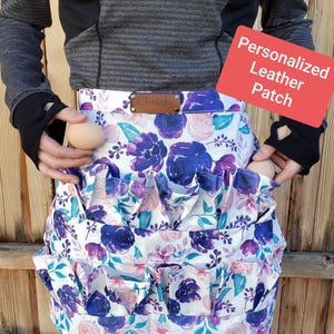 May include: A floral egg-gathering apron with a white background and purple and blue flower pattern. The apron has multiple pockets and a brown leather patch with the name "Christy". The image also includes a red sign that says "Personalized Leather Patch".