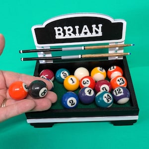 May include: A black wooden billiard rack with a green felt base containing fifteen numbered pool balls and two miniature pool cues. The top of the rack features a black sign with the white text BRIAN. Two pool balls are held in the foreground.