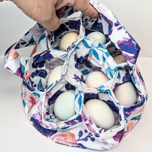 May include: A floral egg carrier with a white background. The carrier is filled with white and light blue eggs. The fabric design includes purple, pink, and blue flowers and leaves. The carrier has a handle and is held by a hand.