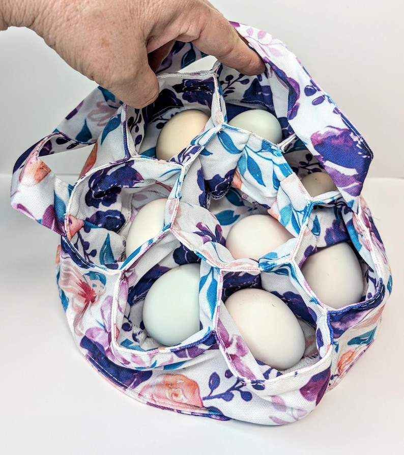 May include: A floral egg carrier with a white background. The carrier is filled with white and light blue eggs. The fabric design includes purple, pink, and blue flowers and leaves. The carrier has a handle and is held by a hand.