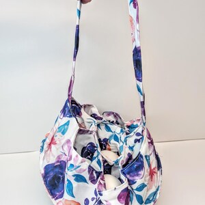 May include: A floral patterned bag with a drawstring closure and a handle. The bag has a white base with purple, blue, and pink flower designs. The bag is designed to hold eggs.