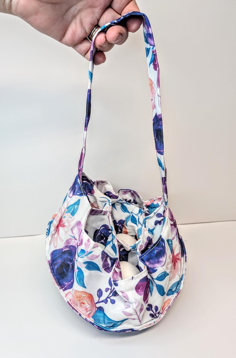 May include: A floral patterned bag with a drawstring closure and a handle. The bag has a white base with purple, blue, and pink flower designs. The bag is designed to hold eggs.