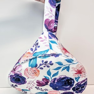 May include: A floral bag with a handle, held up. The bag has a white background with a pattern of purple, pink, and blue flowers and leaves. The handle is made from the same floral fabric as the bag.