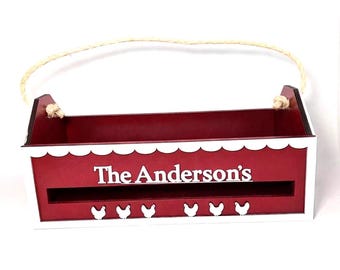 Puede incluir: Una caja rectangular de madera roja con un borde superior festoneado blanco y un asa de cuerda de yute. El frente presenta el texto blanco The Anderson's sobre una ranura horizontal y seis pequeñas siluetas de pollos blancas.