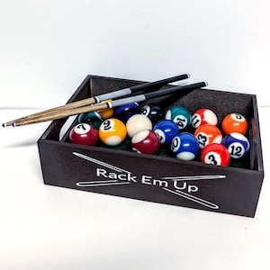May include: A dark wooden rectangular box containing a set of colorful numbered billiard balls and two pool cues resting on top. The front of the box features the white text Rack Em Up with a graphic of two crossed pool cues.