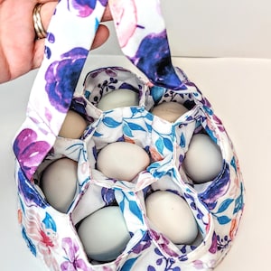 May include: A floral egg gathering basket with a handle, filled with eggs. The fabric features a white background with purple, blue, and pink floral patterns. The basket is designed to hold eggs securely.