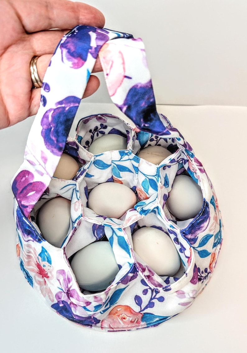 May include: A floral egg gathering basket with a handle, filled with eggs. The fabric features a white background with purple, blue, and pink floral patterns. The basket is designed to hold eggs securely.
