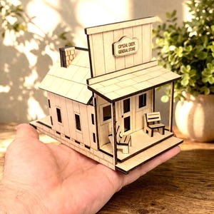 May include: A miniature wooden model of a general store. The model features a sign that reads "Crystal Creek General Store." The building has a porch with two small chairs. The model is held in a hand.