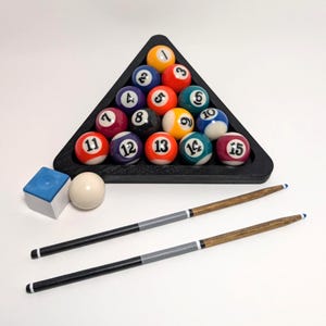 May include: A set of fifteen numbered billiard balls arranged in a black triangular rack, accompanied by a white cue ball, a blue chalk cube, and two wooden pool cues with black and grey handles, all displayed together on a plain white surface.
