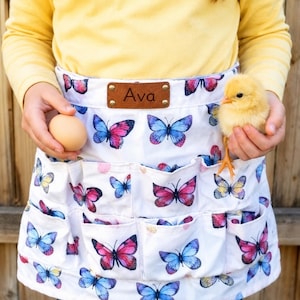 May include: White egg-gathering apron with a butterfly pattern in pink and blue. The apron has multiple pockets and a leather tag that says "Ava". A person is holding an egg and a yellow chick.