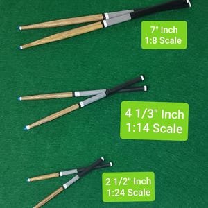 May include: Three pairs of wooden drumsticks in different sizes, with black and grey painted tips. The drumsticks are arranged in a descending order of size, with the largest pair at the top and the smallest pair at the bottom. The image includes text indicating the size and scale of each pair of drumsticks: 7" Inch 1:8 Scale, 4 1/3" Inch 1:14 Scale, and 2 1/2" Inch 1:24 Scale.