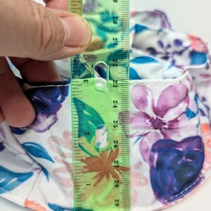 May include: A floral print hat with a ruler showing a measurement of approximately 7.6 cm. The hat features a white background with blue, purple, and pink flower patterns. The ruler is green with black markings.