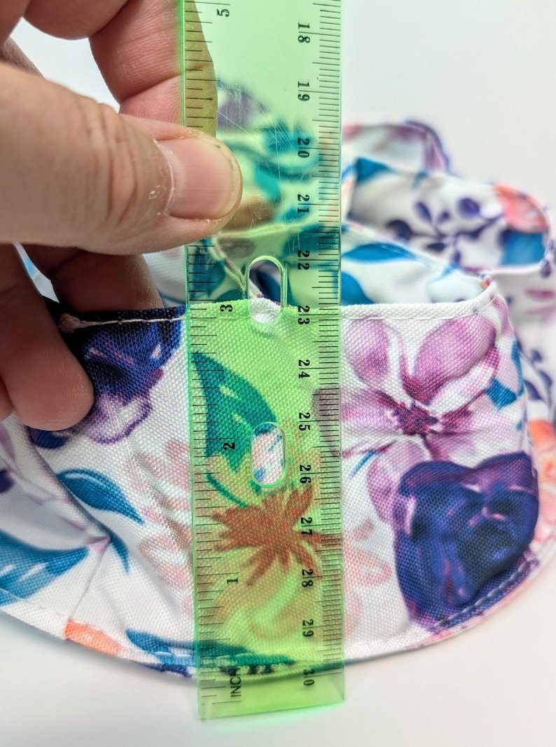 May include: A floral print hat with a ruler showing a measurement of approximately 7.6 cm. The hat features a white background with blue, purple, and pink flower patterns. The ruler is green with black markings.
