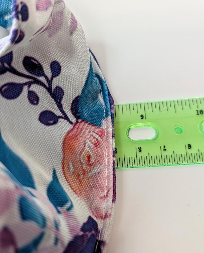 May include: Close-up of a floral patterned fabric with a ruler showing measurements. The fabric features a white background with teal, purple, and pink floral designs. The ruler shows a measurement of approximately 2.5 cm.