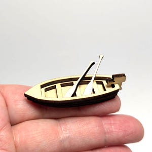 Peut inclure: Un petit bateau en bois avec deux rames et un petit moteur. Le bateau est brun clair avec des bordures brun foncé. Les rames sont brun clair et sont placées à l'intérieur du bateau. Le bateau est tenu dans une main.
