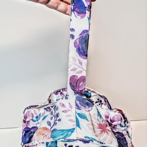 May include: A bag with a floral pattern and a handle. The fabric has a white base with purple, blue, and pink flowers and green leaves. The handle is a long, narrow strip of the same floral fabric.