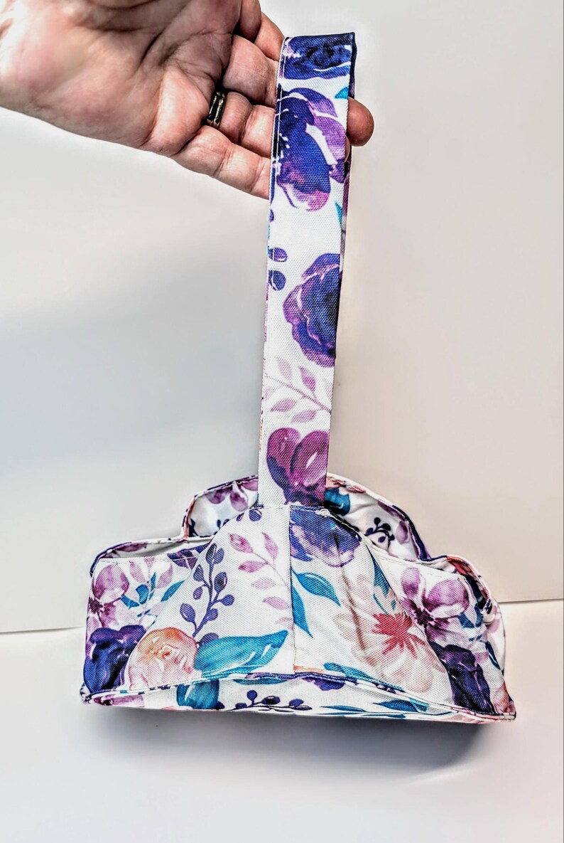 May include: A bag with a floral pattern and a handle. The fabric has a white base with purple, blue, and pink flowers and green leaves. The handle is a long, narrow strip of the same floral fabric.