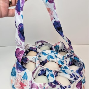 May include: Floral egg gathering basket with a handle, filled with white eggs. The fabric features a white background with purple, blue, and pink floral patterns. The basket has individual compartments for each egg.