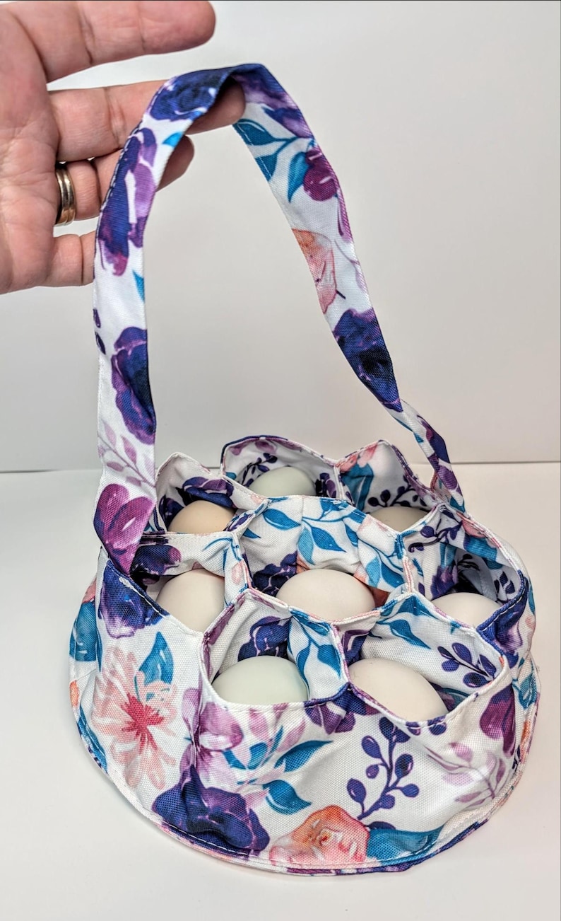 May include: Floral egg gathering basket with a handle, filled with white eggs. The fabric features a white background with purple, blue, and pink floral patterns. The basket has individual compartments for each egg.