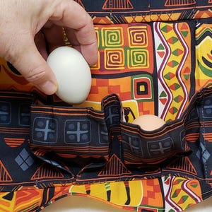 Puede incluir: Un porta huevos de tela negra con un diseño de estampado africano colorido. El porta huevos tiene varios bolsillos para guardar huevos. Dos huevos blancos son visibles en los bolsillos.