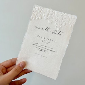 May include: A cream-colored wedding invitation with embossed floral detailing at the top. The text "save the date" is written in elegant script, followed by the names "SAM & PENNY" and wedding details. The paper has a textured, torn edge.