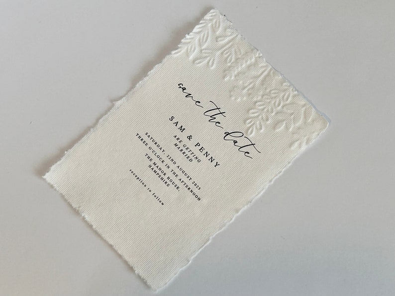 May include: Off-white textured paper invitation with a torn edge. The top right corner features an embossed floral design. The text reads "save the date" and includes wedding details: names, date, time, and location.