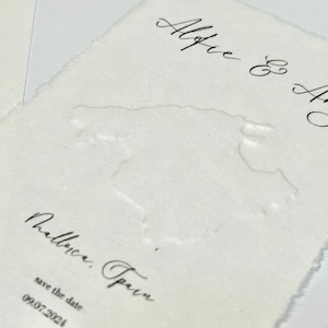 Könnte beinhalten: Off-white strukturiertes Papier mit den Namen "Alfie & Amy" in eleganter Schrift. Darunter sind die Worte "Mallorca, Spain" und das Datum "09.07.2024" gedruckt. Eine subtile geprägte Karte von Mallorca ist sichtbar.
