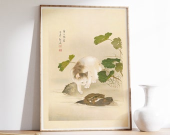 Puede incluir: Una impresión japonesa en madera enmarcada que representa un gato blanco mirando hacia abajo a una rana en una roca. La impresión está sobre un fondo crema con hojas verdes y texto japonés en la esquina superior izquierda.