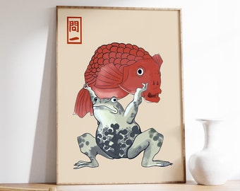 Op de afbeelding: Ingelijste kunstdruk met een kikker die een grote rode vis omhoog houdt. De kikker is grijs met zwarte vlekken en de vis heeft een gedetailleerd schubbenpatroon. Het kunstwerk heeft een beige achtergrond en een rood en zwart Japans karakter.