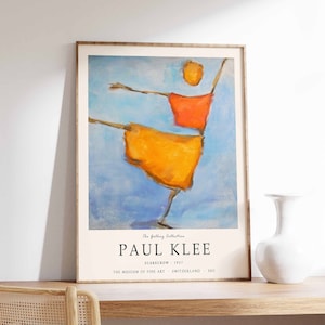 May include: A framed art print of Paul Klee's "Scarecrow" from 1927. The artwork features an abstract figure in orange and yellow against a blue background. The print includes the artist's name and the museum details.