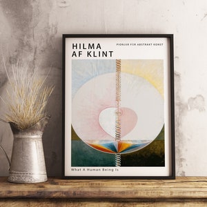 Puede incluir: Un cuadro enmarcado con un diseño abstracto colorido, con el texto "HILMA AF KLINT" y "What A Human Being Is." Una jarra de metal con hierba seca está sobre una estantería de madera.