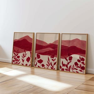 May include: Three framed art prints featuring a watercolor landscape with red mountains and floral accents. The artwork has a beige background and is framed in gold. The floral elements are red.