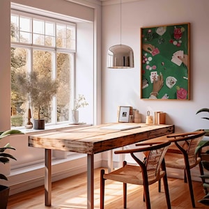 May include: A bright, airy room with a wooden table and two chairs. A framed artwork depicting a poker game hangs on the wall. A large window offers a view of trees. A silver pendant light hangs above the table.