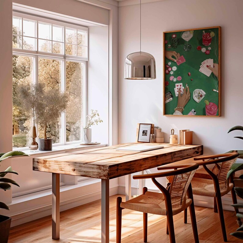 May include: A bright, airy room with a wooden table and two chairs. A framed artwork depicting a poker game hangs on the wall. A large window offers a view of trees. A silver pendant light hangs above the table.